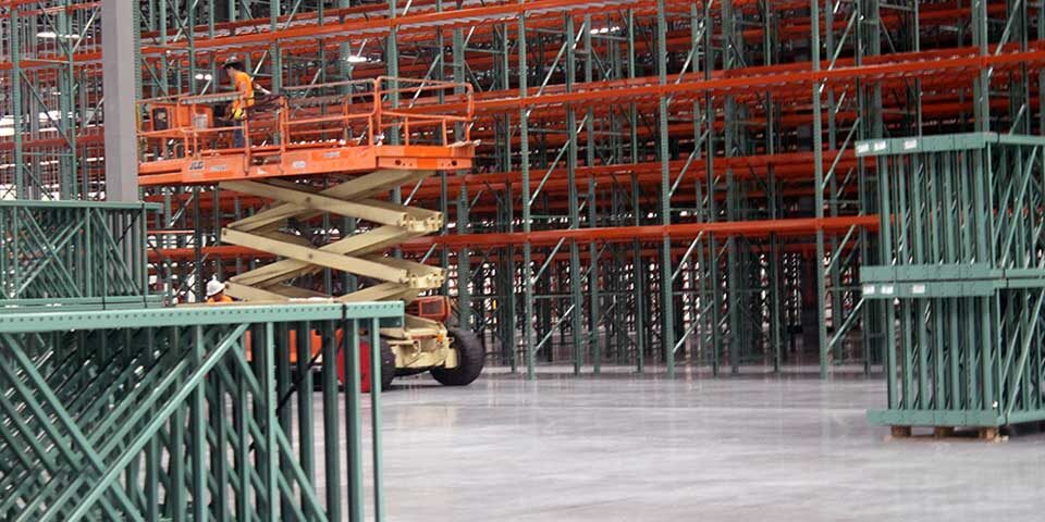 A warehouse foreman wearing a hi-vis jacket and a hard hat while operating a scissor lift in an open warehouse filled with pallet storage shelves from floor to ceiling.