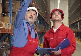 Two warehouse workers, one of which is a foreman, both wearing hi-vis jackets and hard hats are discussing safety requirements and pointing to the top of a storage rack system.